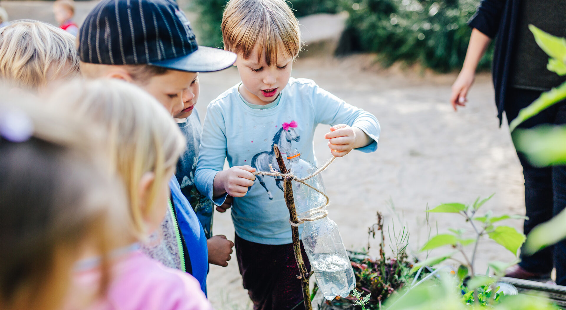 Ein Kind versucht, eine Plastikflasche mit Wasser an einem Stock in einem Hochbeet festzubinden, andere Kinder schauen dabei zu