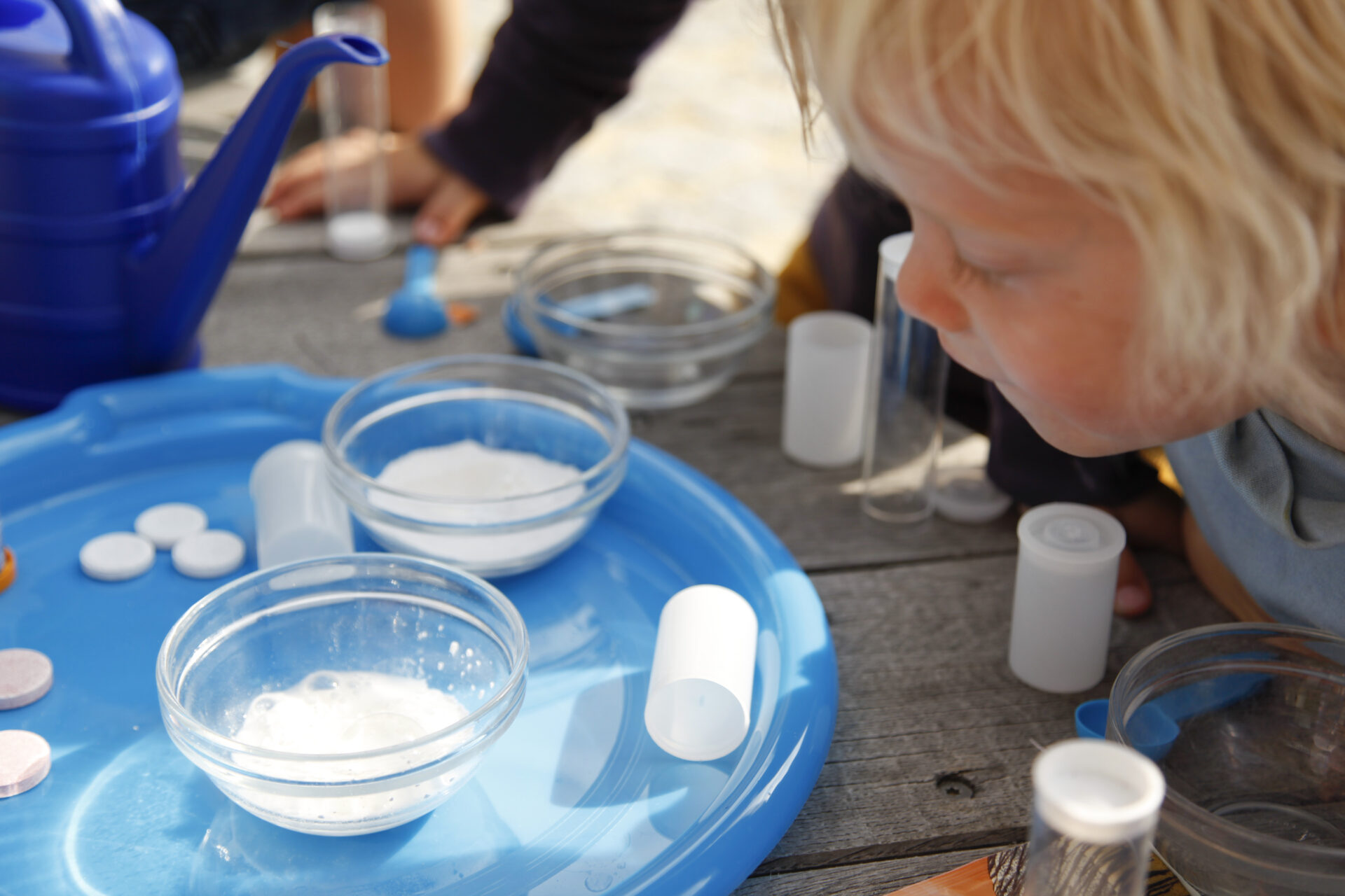 Ein Kind blickt auf ein Tablett auf dem kleine Glasschalen, Filmdosen und Brausetabletten liegen. In einer Glaschale befindet sich eine sprudelnde Masse.